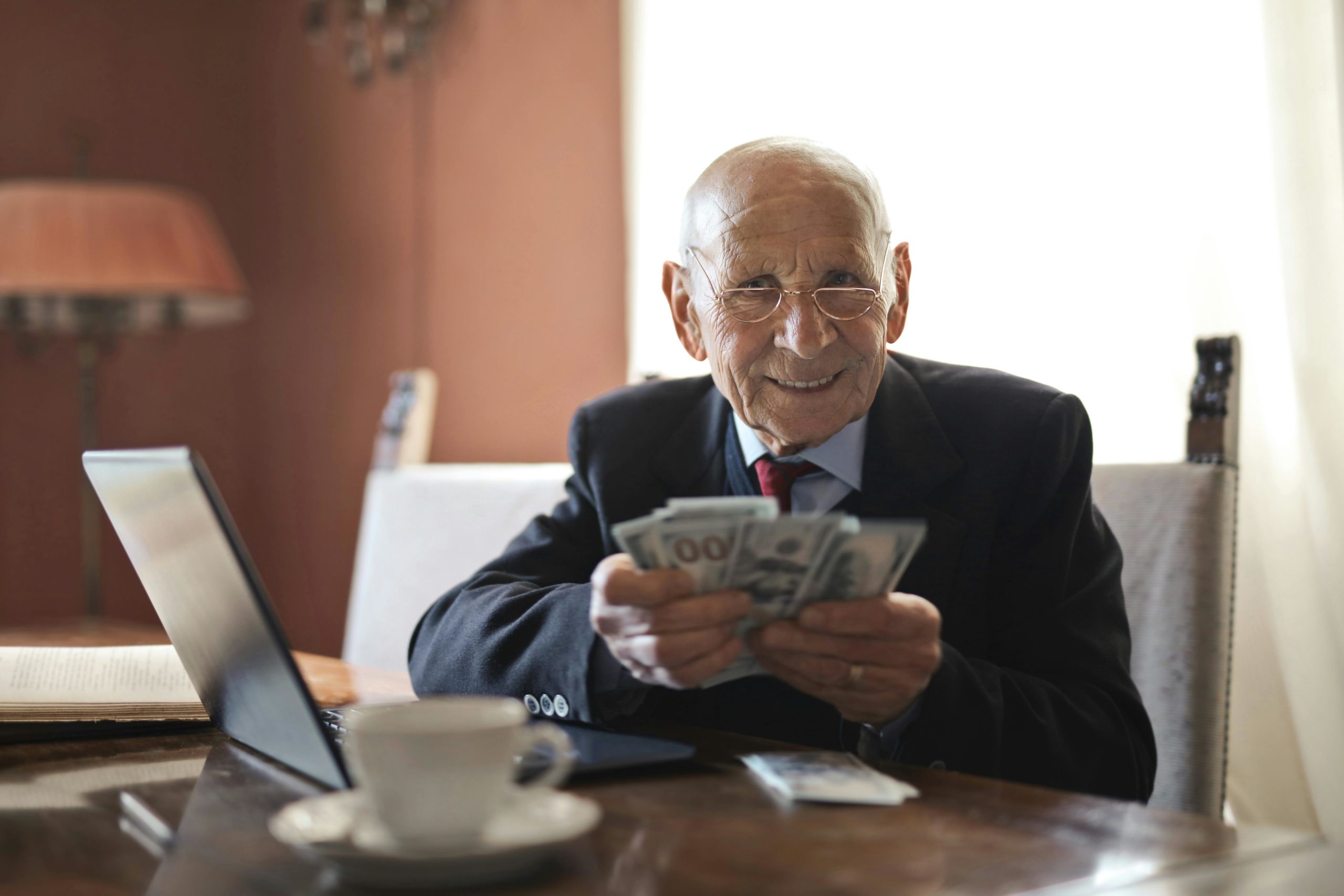 Selbstbewusster älterer Geschäftsmann, Der Geld In Den Händen Hält, Während Er Am Tisch Nahe Laptop Sitzt