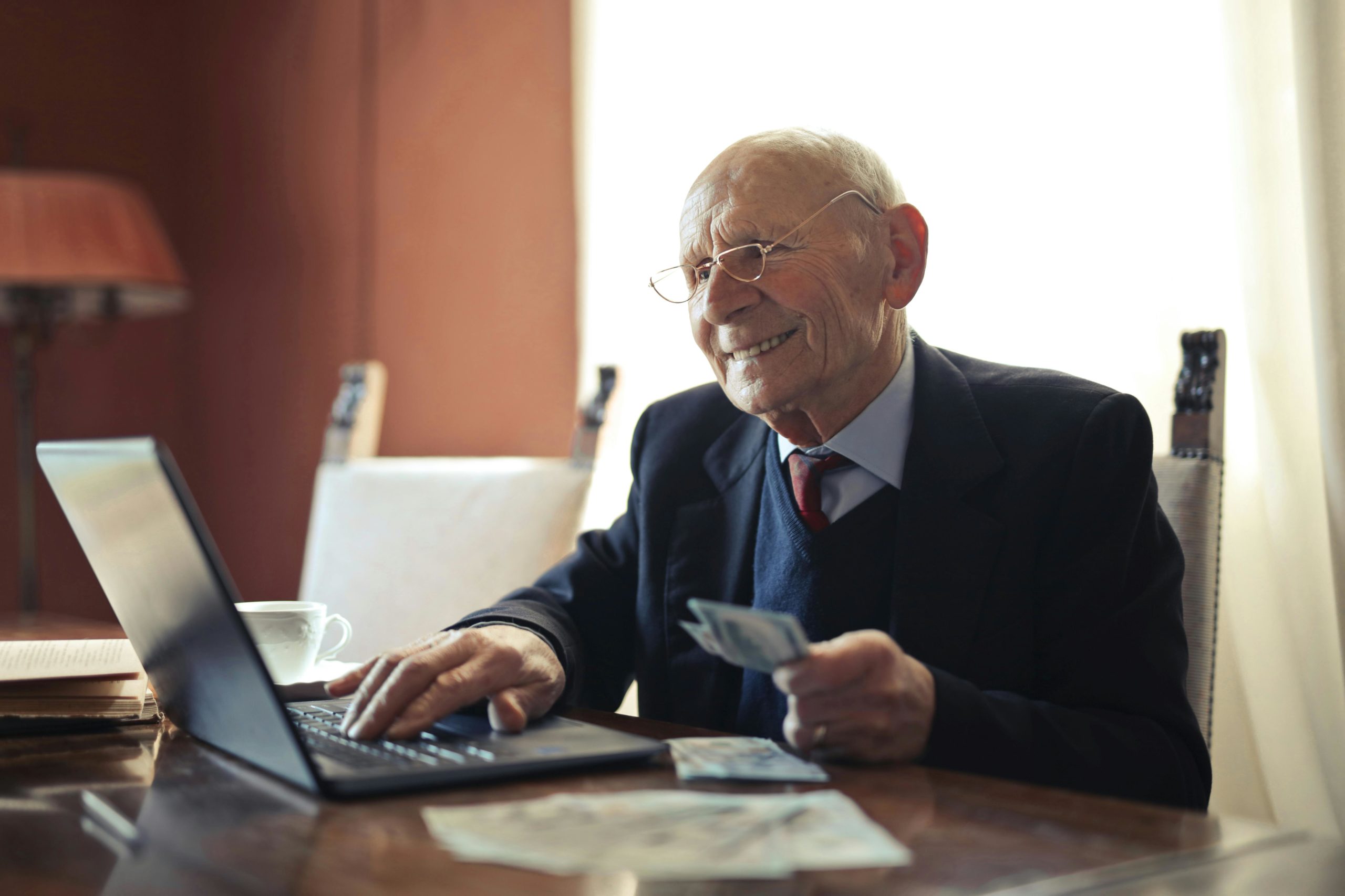 Positiver älterer Geschäftsmann, Der Auf Laptop Schreibt, Während Er Geld In Der Hand Hält