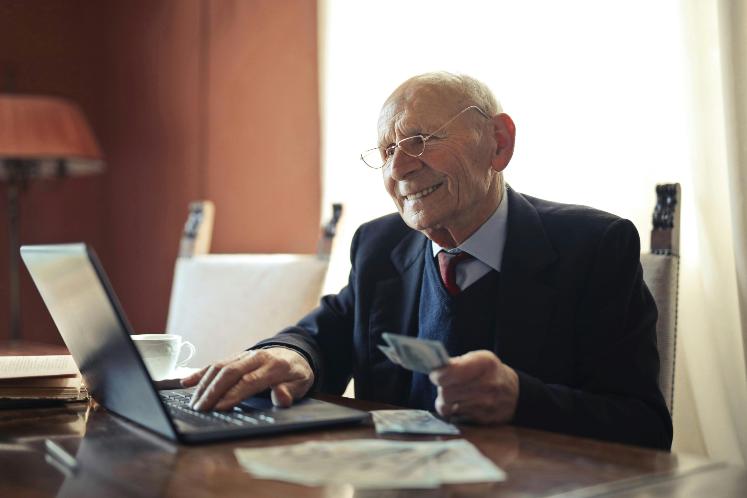 Positiver älterer Geschäftsmann, Der Auf Laptop Schreibt, Während Er Geld In Der Hand Hält