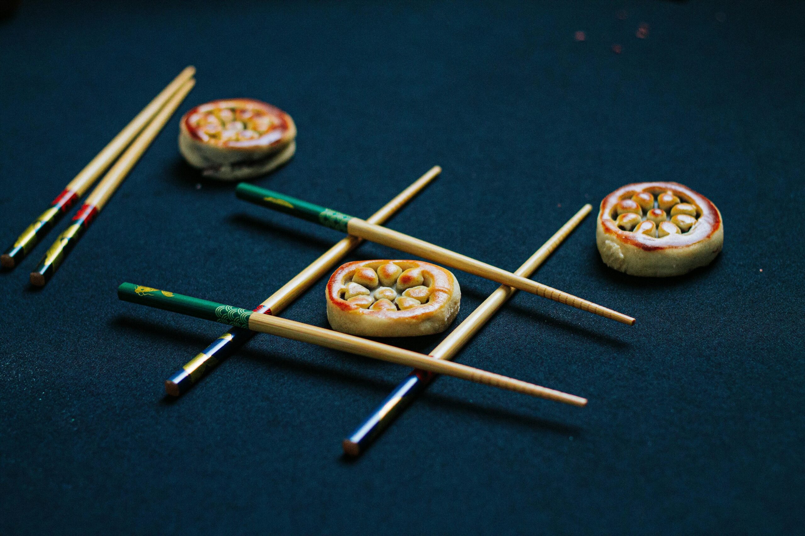 Entdecken Sie ein spielerisches Tic-Tac-Toe-Setup mit bunten Essstäbchen und Mochi auf einer dunklen Oberfläche.
