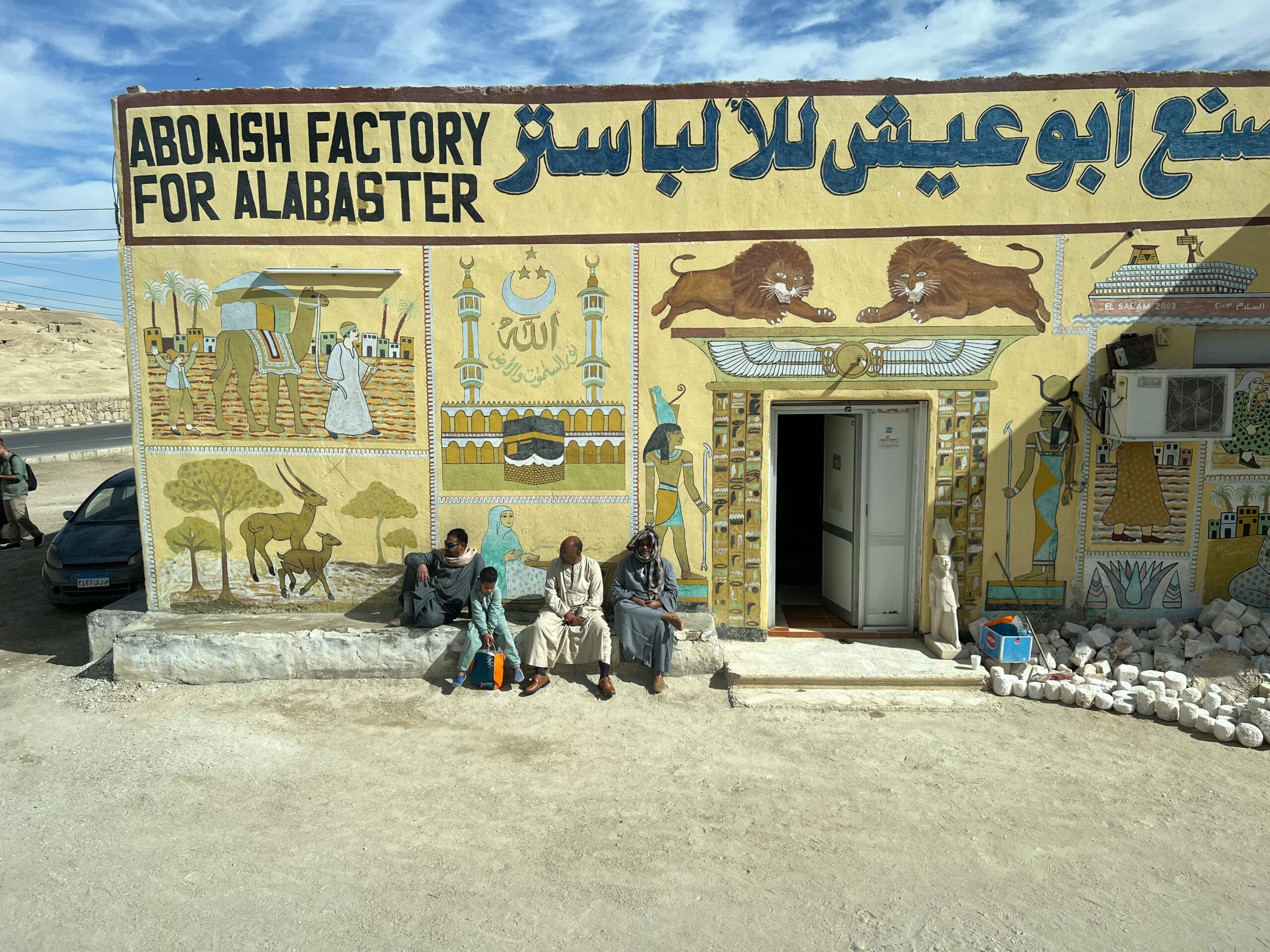 Drei Männer sitzen vor der Aboaish Alabaster Factory und bewundern die farbenfrohen Wandgemälde in einer sonnigen Umgebung im Freien.