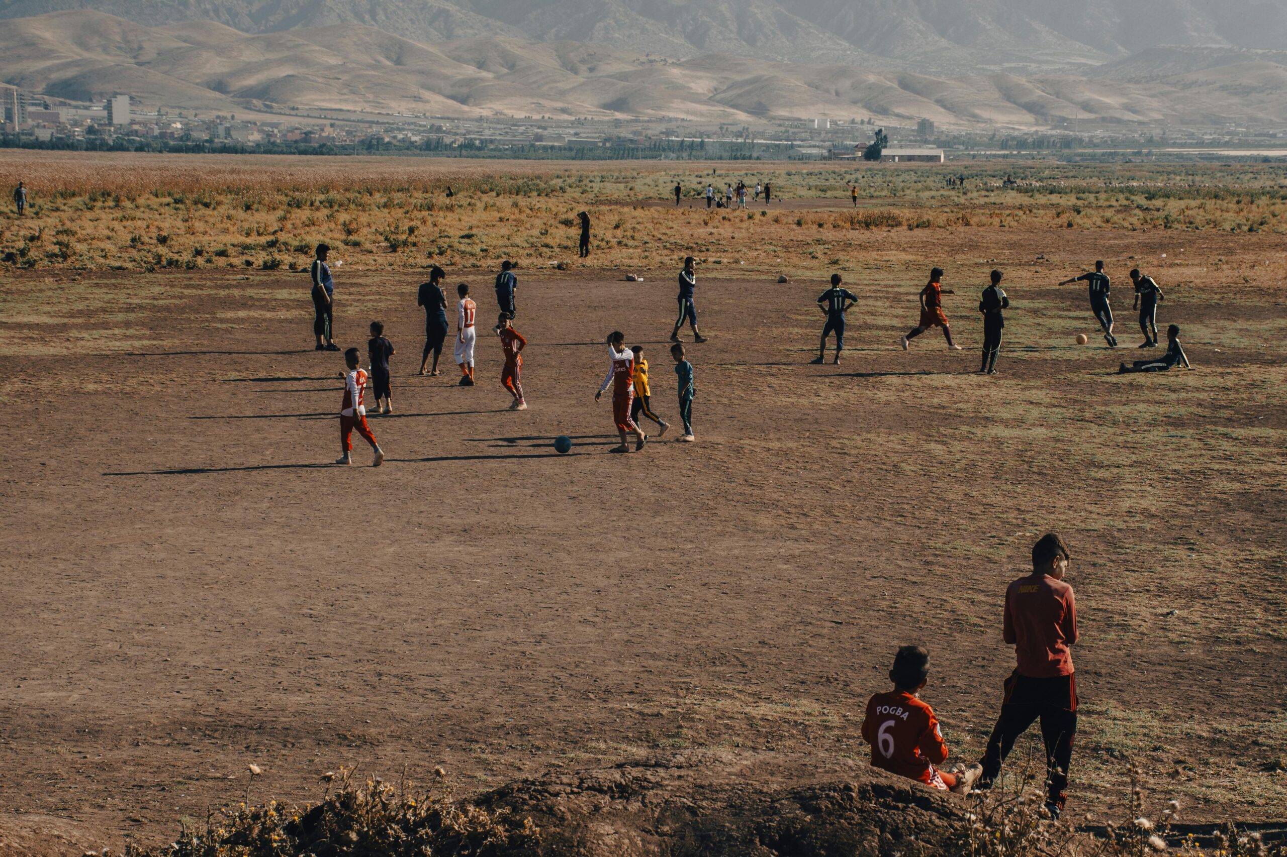 Eine Gruppe Kinder spielt Fußball auf einem Feld im ländlichen Raum mit Bergen im Hintergrund. Die Szene fängt eine warme, stimmungsvolle Atmosphäre ein.